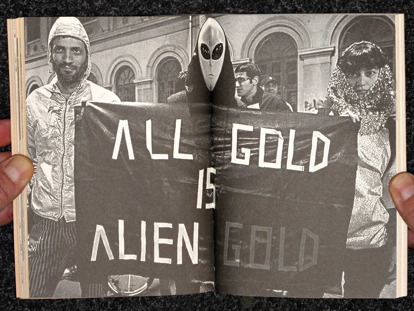 A black-and-white spread of people holding a banner that reads "All gold is alien gold"