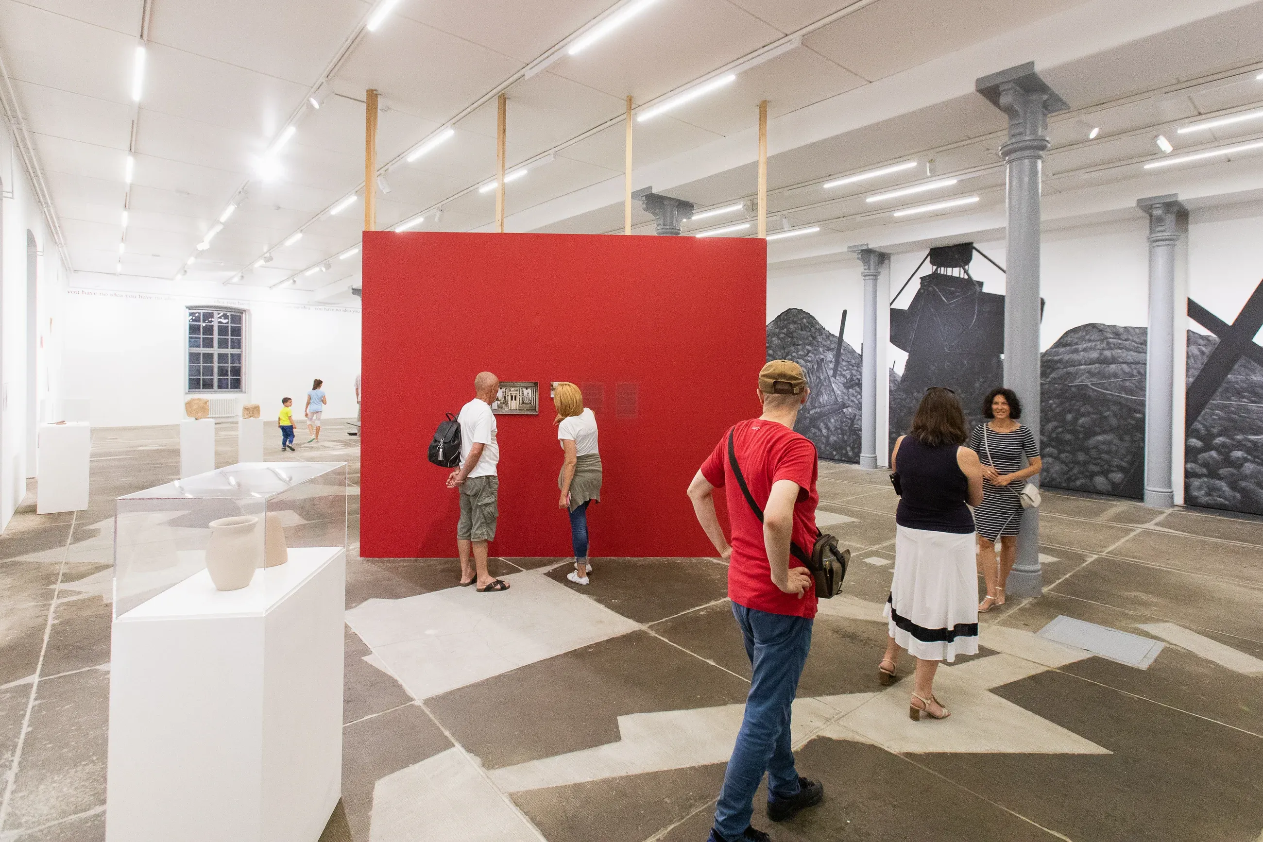 A red free-standing wall in a large industrial hall, with visitors in front of it